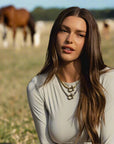 A woman wearing a white long-sleeve top and blue denim shorts with a Stellar Cheval necklace sitting outdoors with a fence and trees and horses in the background.
