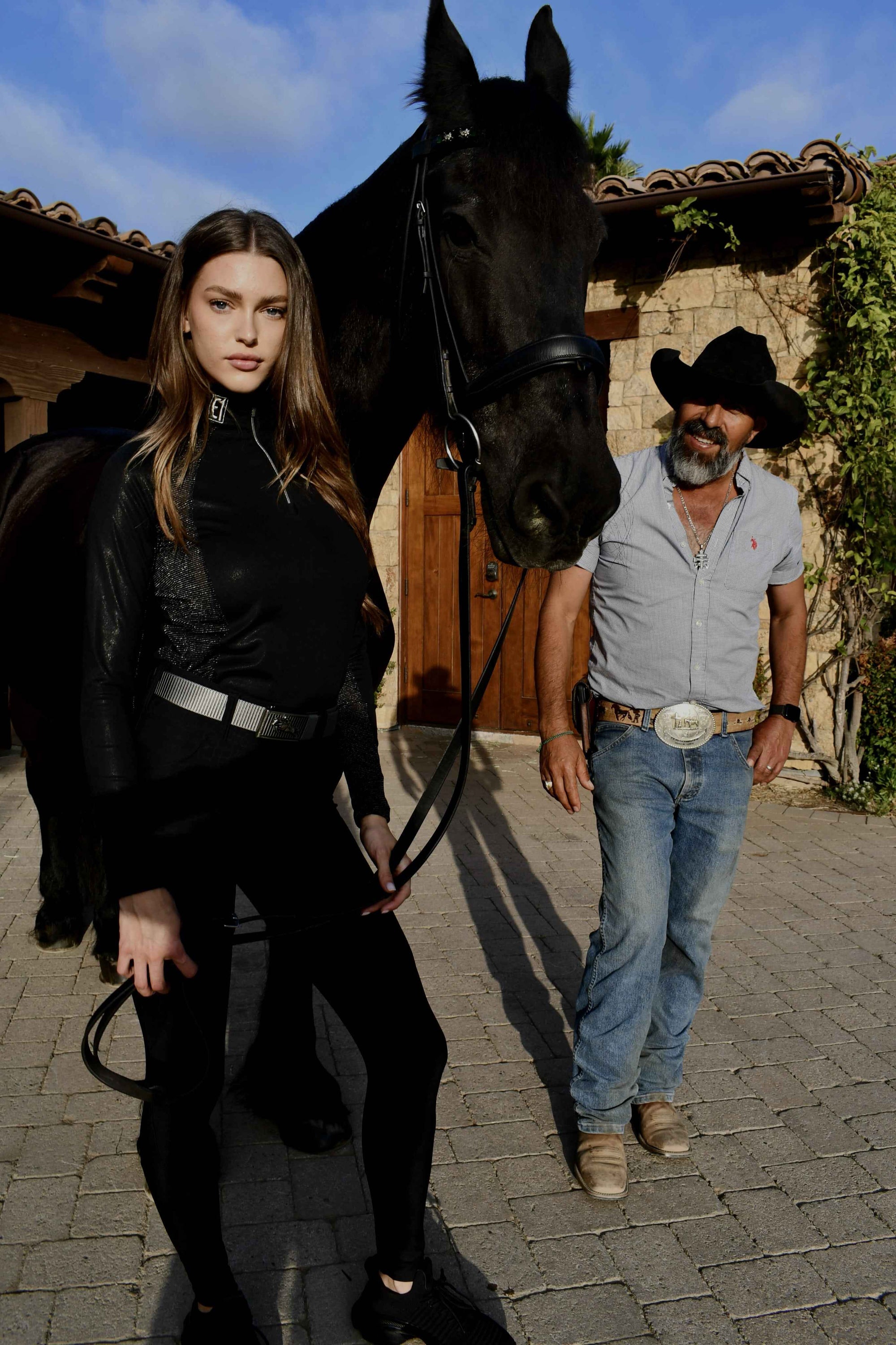 Woman in equestrian black outfit holding a horse's bridle, man in cowboy hat and jeans walking beside her.